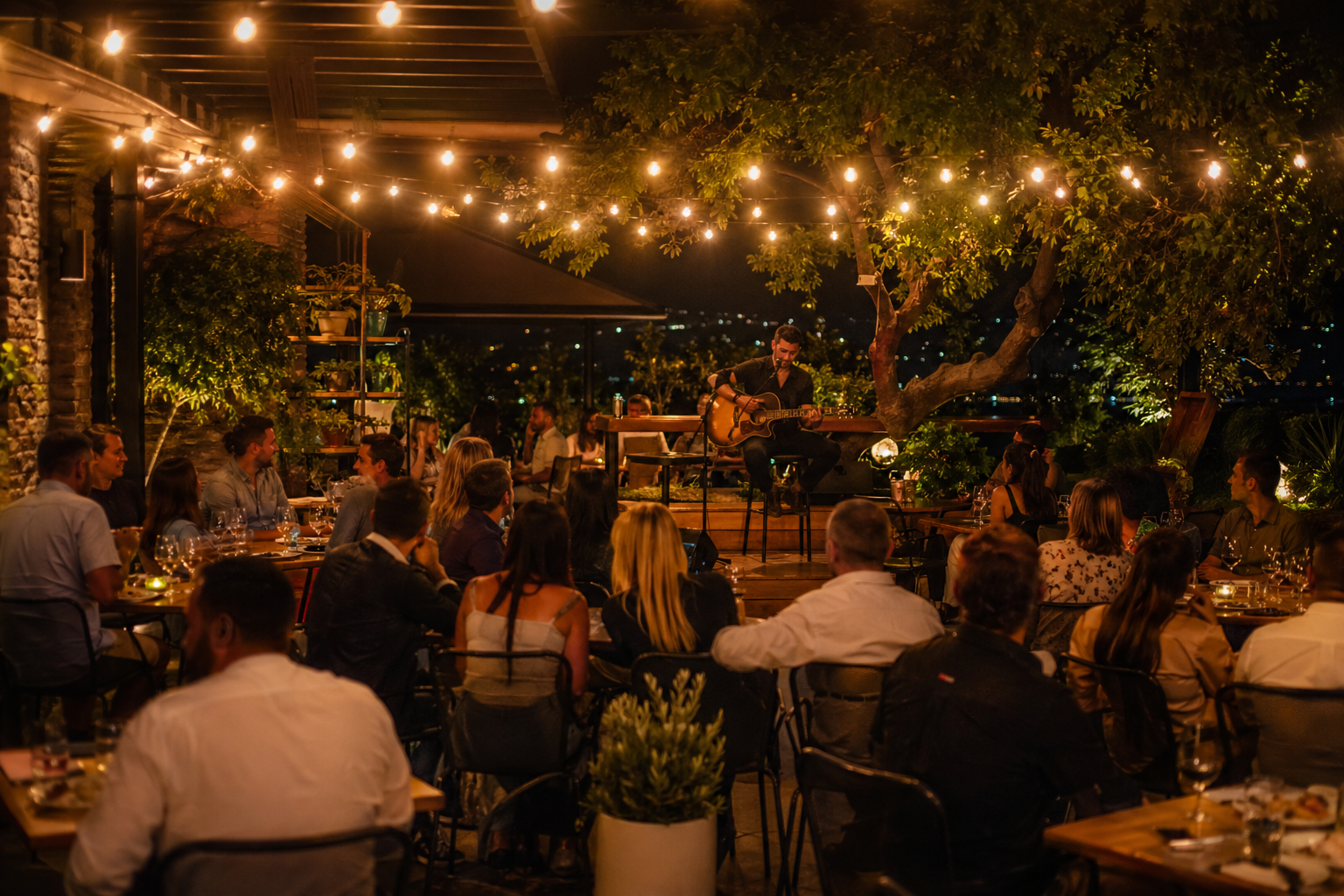 Outdoor restaurant patio at night with string lights and solo acoustic guitarist performing for a full crowd